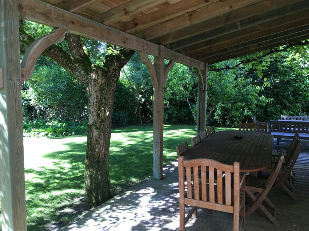 Longue terrasse en bois sous pergola, ombragée par un grand arbre. Table et chaises pour un repas convivial dans le jardin verdoyant.