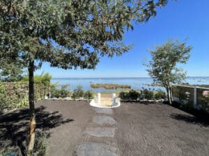 Aménagement de jardin en bord de mer. Terre labourée et pas japonais mènent à une vaste lagune bleue parsemée de voiliers sous un ciel clair.