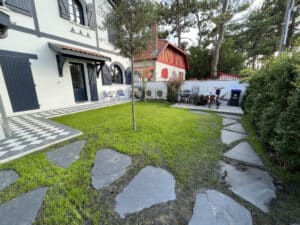 Façade blanche et grise d'une maison de style Atlantique avec volets bleus. Patio à carreaux damier, pelouse neuve et chemin d'ardoise.
