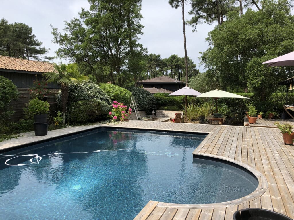 Piscine, terrasse en bois et jardin luxuriant pour l'été. Piscine extérieure bleu foncé bordée par une terrasse en bois clair. Jardin luxuriant, parasols et hydrangeas roses créent un espace de détente.