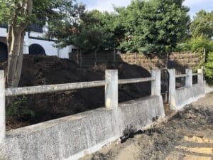 Grand tas de terre végétale noire devant une villa blanche et des arbres. Clôture basse en béton rugueux près d'un sol sablonneux.